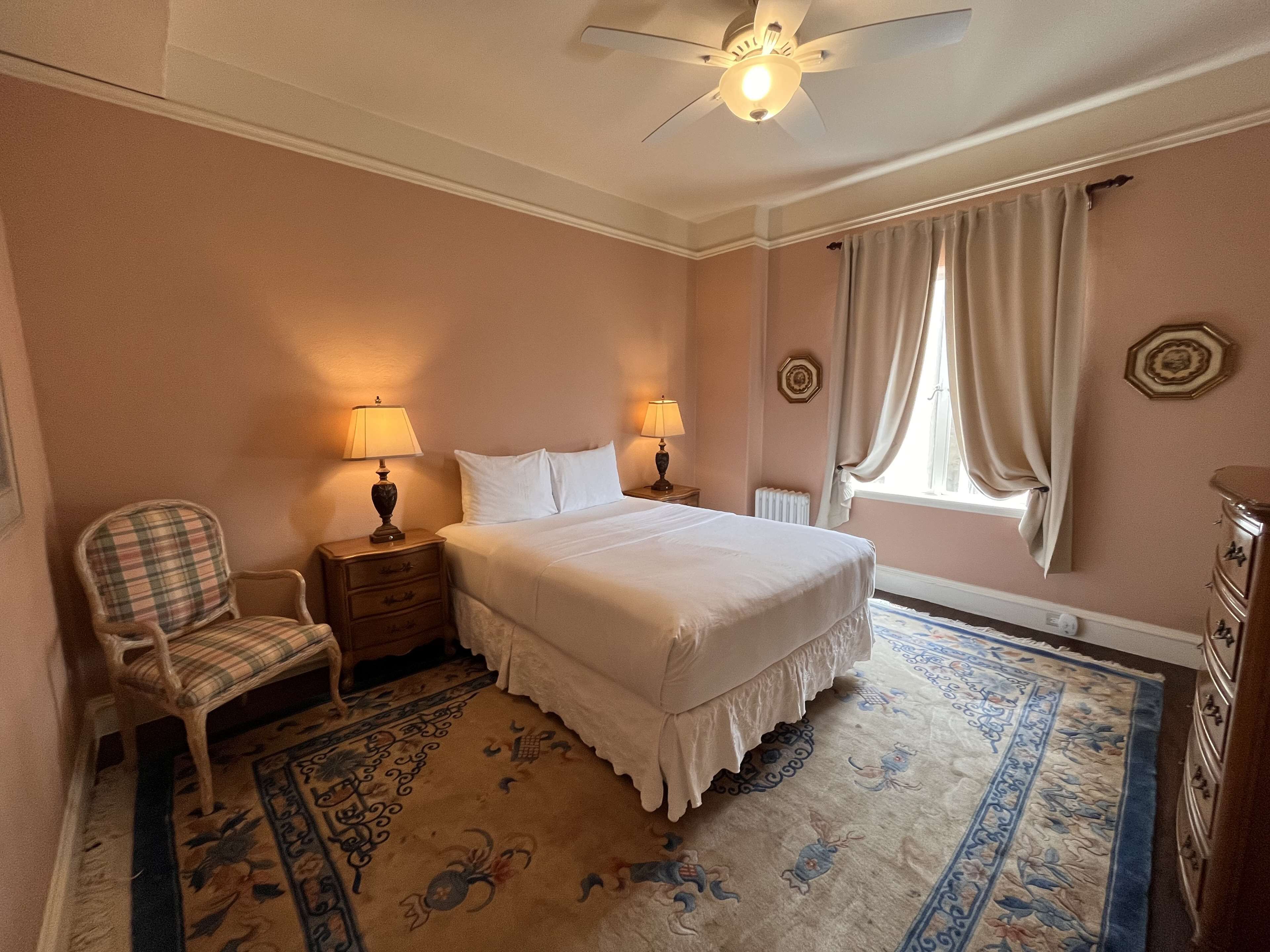 Un dormitorio con cama, dos mesitas de noche con lámparas, una silla, un ventilador de techo, una ventana con cortinas y una alfombra decorativa.