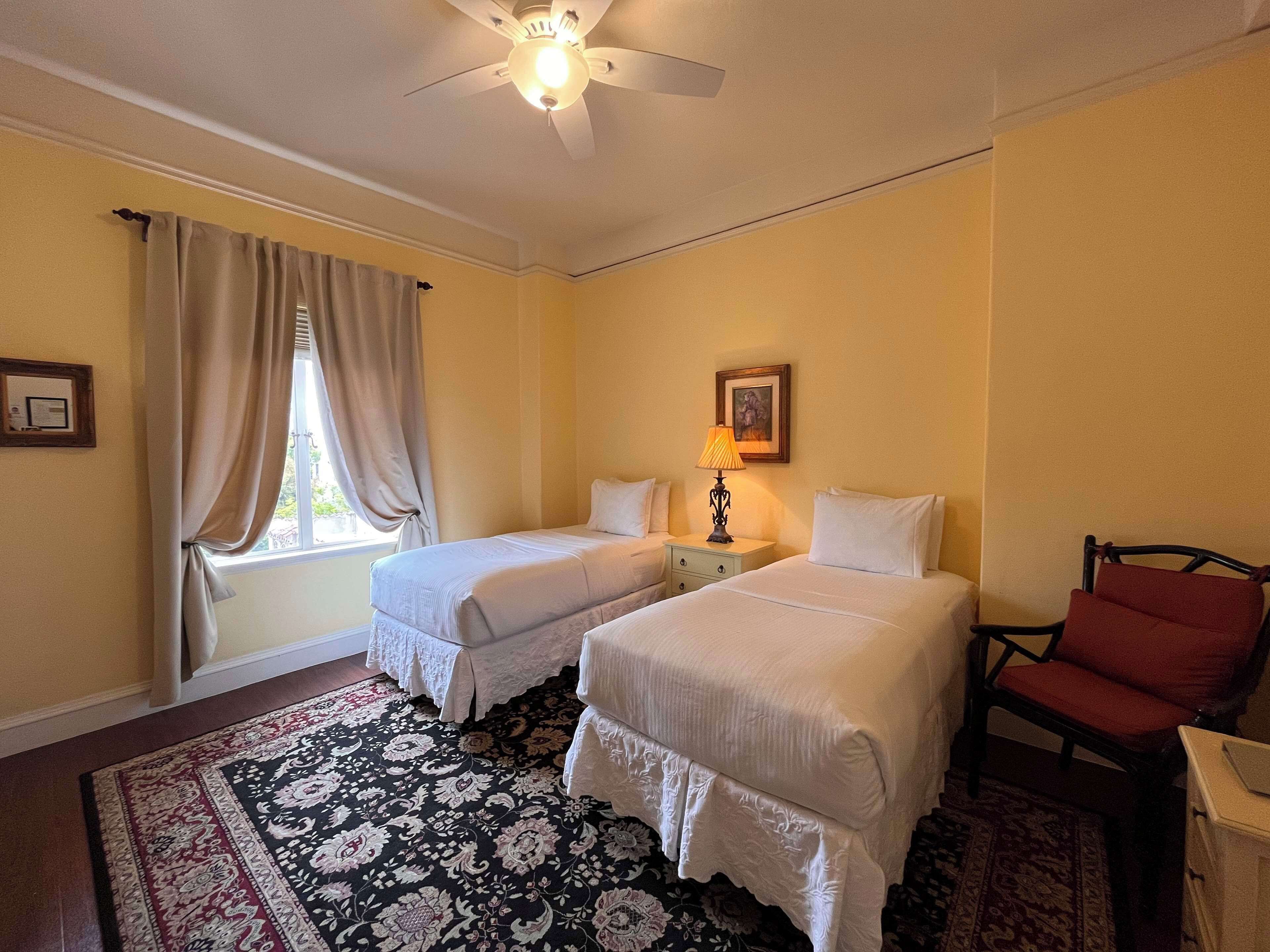 Une chambre d’hôtel avec deux lits, un bureau, une chaise, un ventilateur de plafond et une fenêtre avec des rideaux.