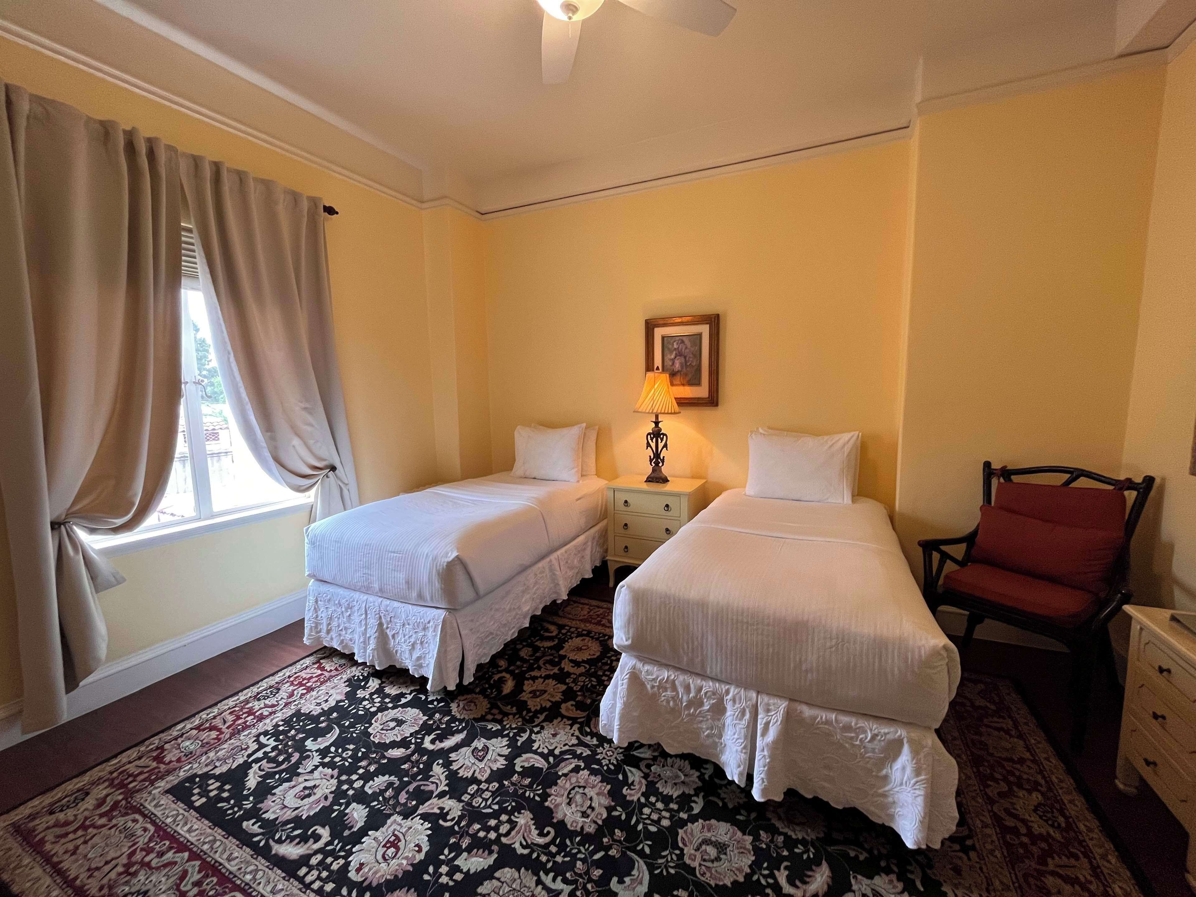 A hotel room with two beds, a nightstand with a lamp, a chair, and a patterned rug.