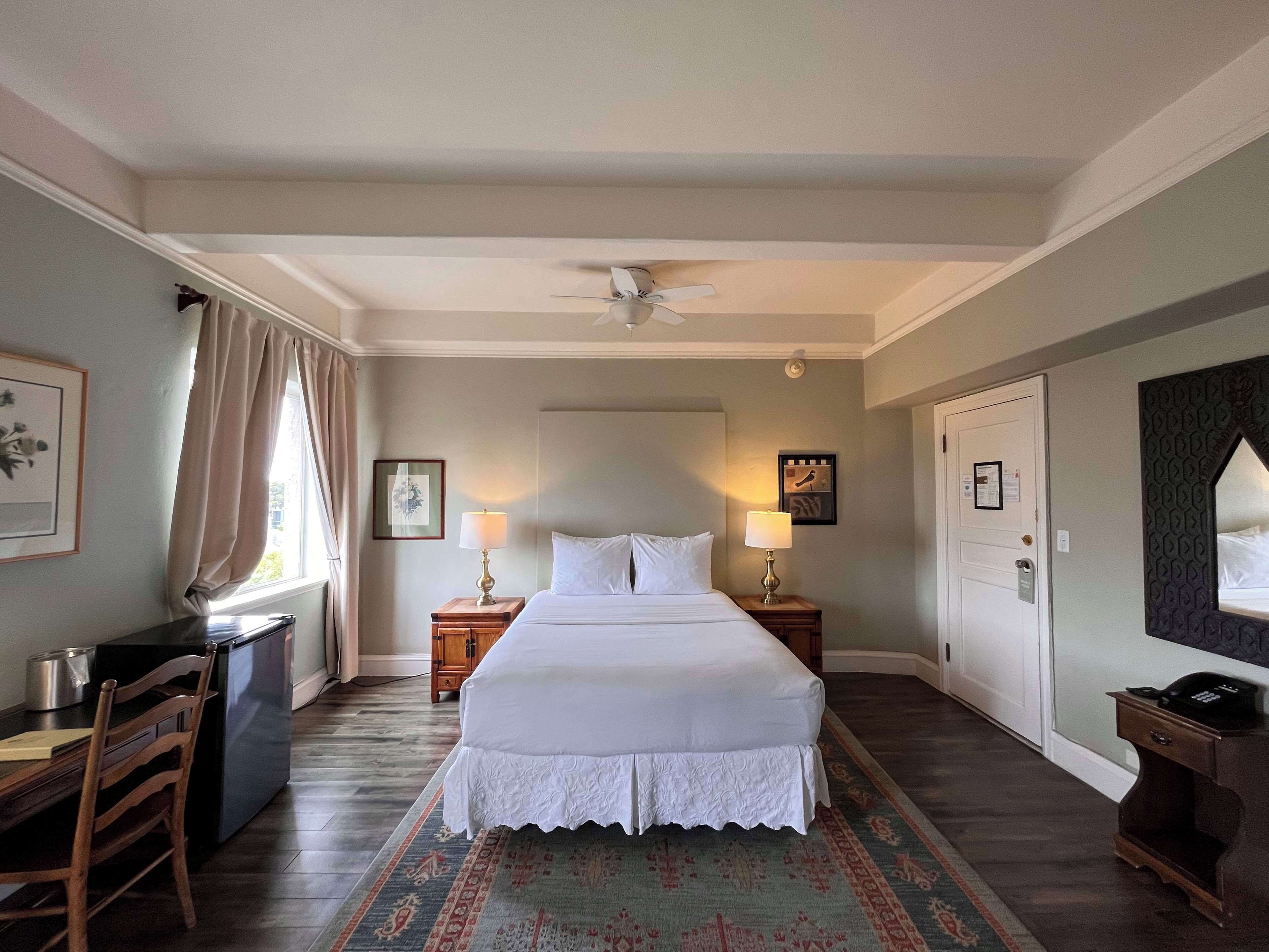 Una habitación de hotel con una cama, dos mesitas de noche con lámparas, un escritorio con silla, un ventilador de techo y una alfombra estampada.