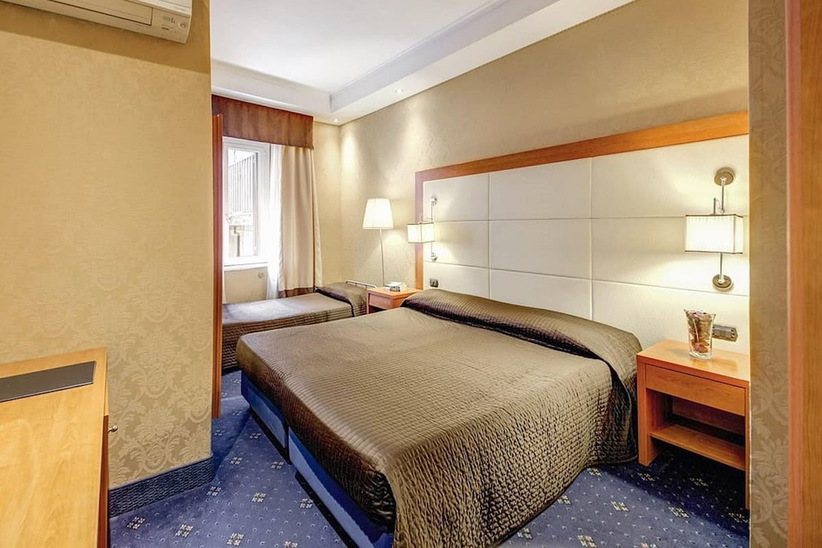 Hotellihuone, jossa on suuri sänky, yöpöytä lampun kanssa, pieni pöytä kannullisineen ja televisio.
