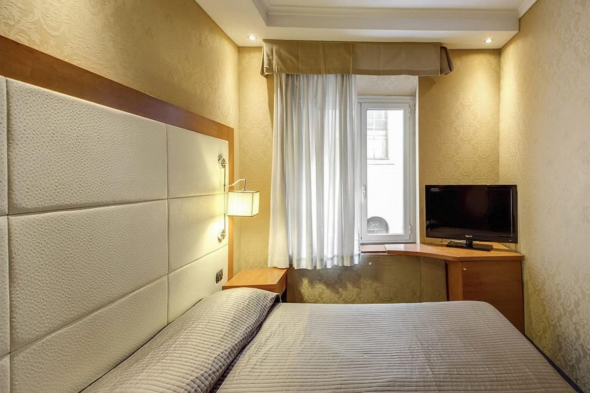Hotellihuone, jossa on sänky, työpöytä, jossa on televisio, ikkuna verhoilla ja seinään kiinnitetty lamppu.