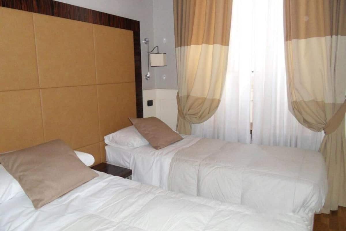 Hotellihuone, jossa on kaksi sänkyä, beigenväriset verhot ja seinään kiinnitetty valaisin.