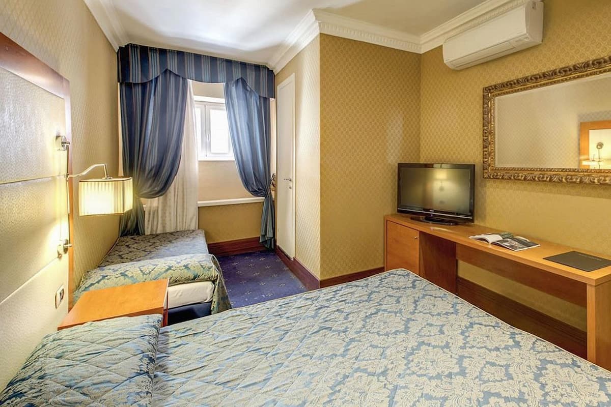 Hotellihuone, jossa on kaksi sänkyä, televisio, työpöytä, peili ja ikkuna, jossa on verhot.