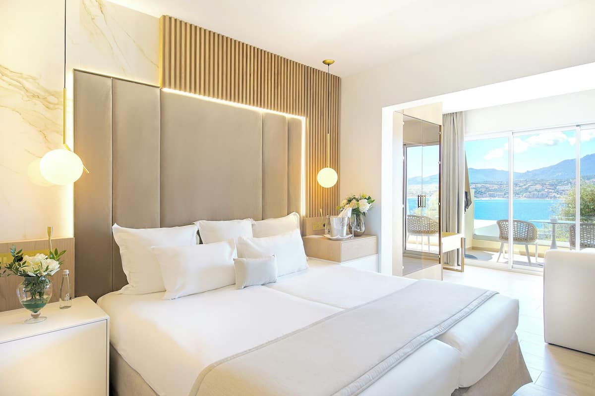 A modern hotel room with a large bed, a nightstand, a vase with flowers, and a view of the sea.