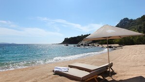 Une plage avec deux chaises longues placées sous un auvent, une serviette sur l’une des chaises, et une vue dégagée sur la mer et les collines lointaines.