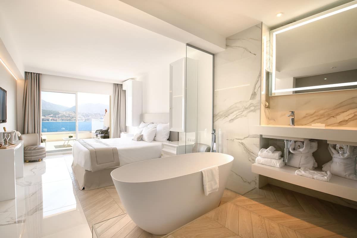 A modern hotel room with a large bathtub, a bed, a vanity with a mirror, and a view of the city.