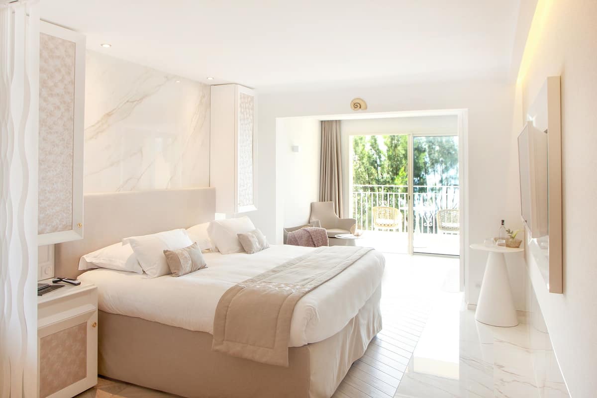 A modern hotel room with a large bed, a small table, and a balcony with a view of greenery.