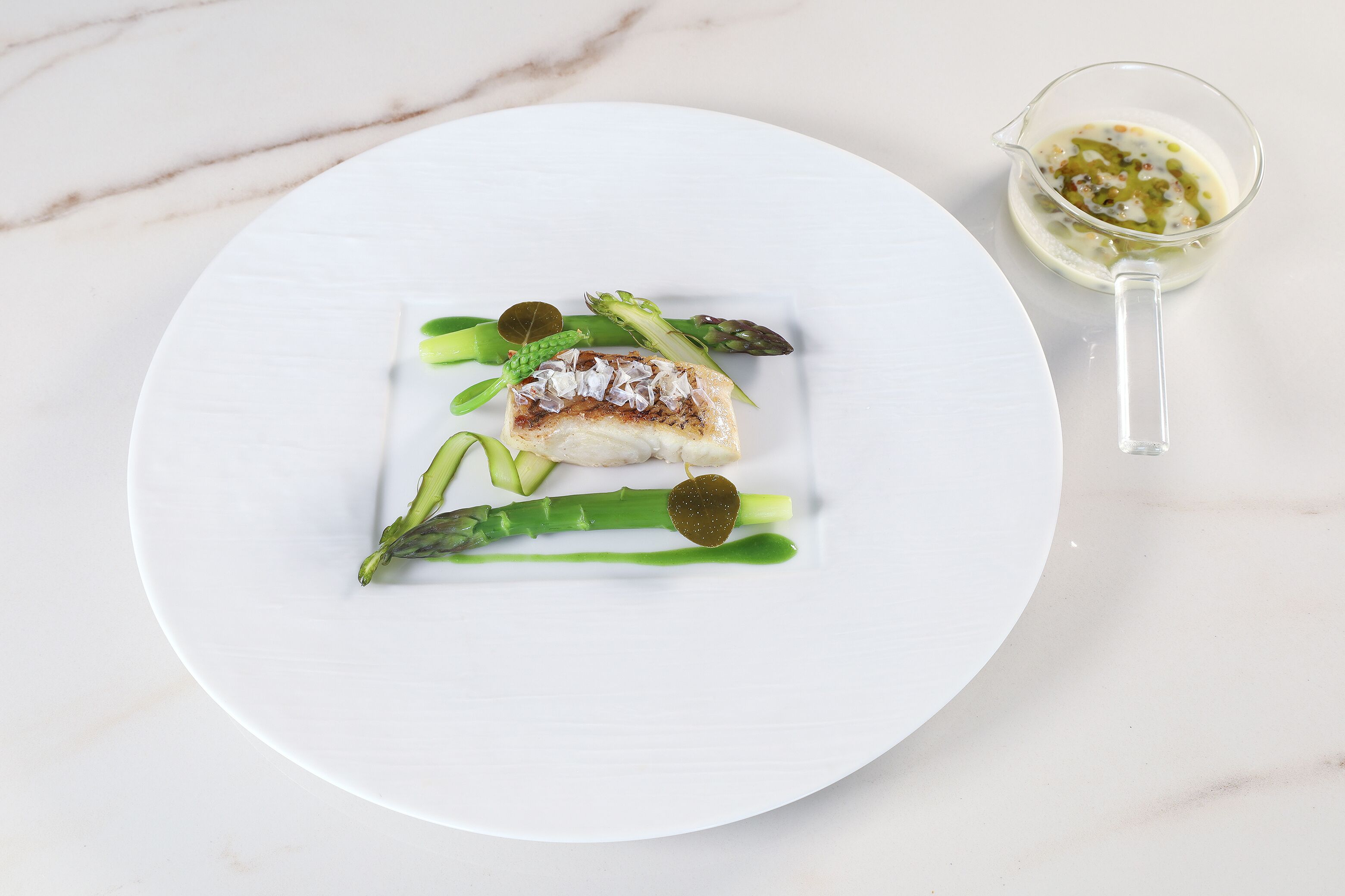 A white plate with a piece of fish, asparagus, and a sauce on a marble surface.
