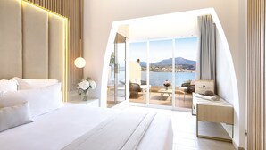 Une chambre moderne avec un grand lit, un bureau et un balcon offrant une vue sur la mer et les montagnes.