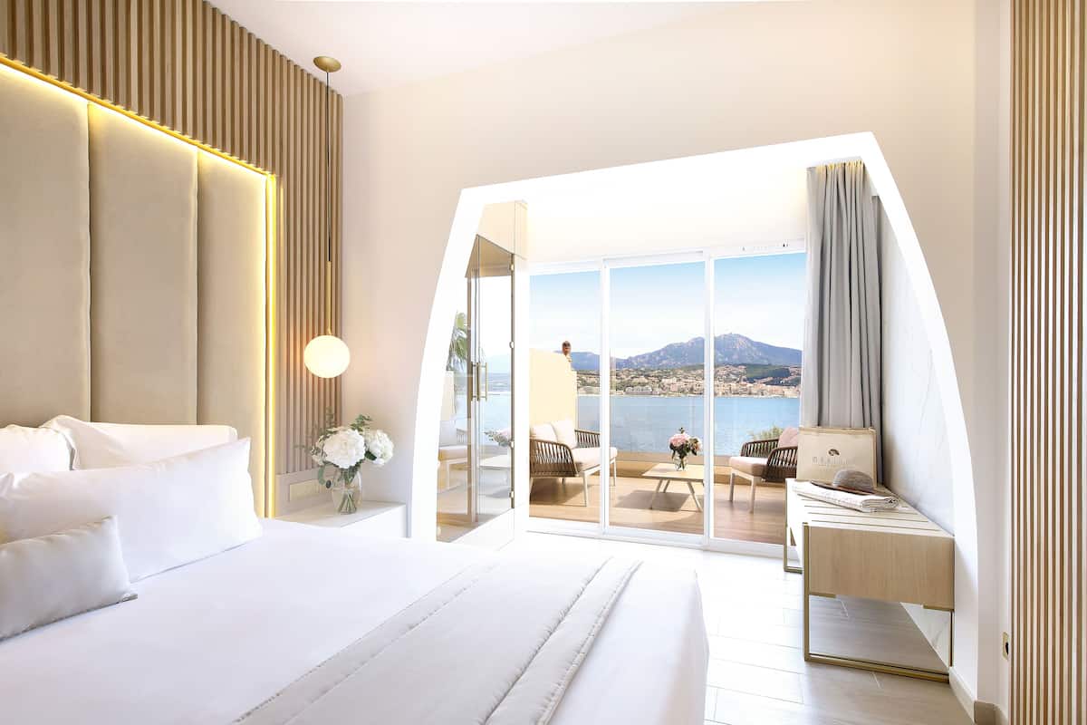 A modern bedroom with a large bed, a desk, and a balcony with a view of the sea and mountains.