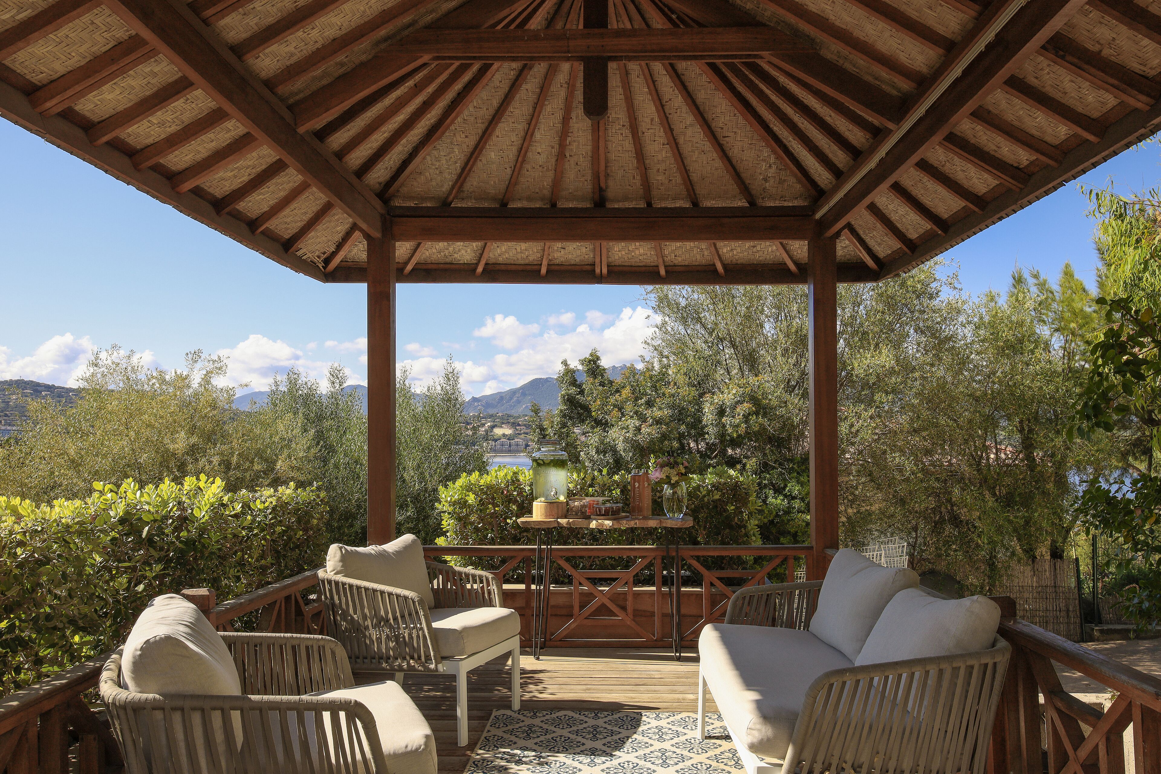 A covered outdoor seating area with wicker furniture and a table with drinks, overlooking a scenic landscape.