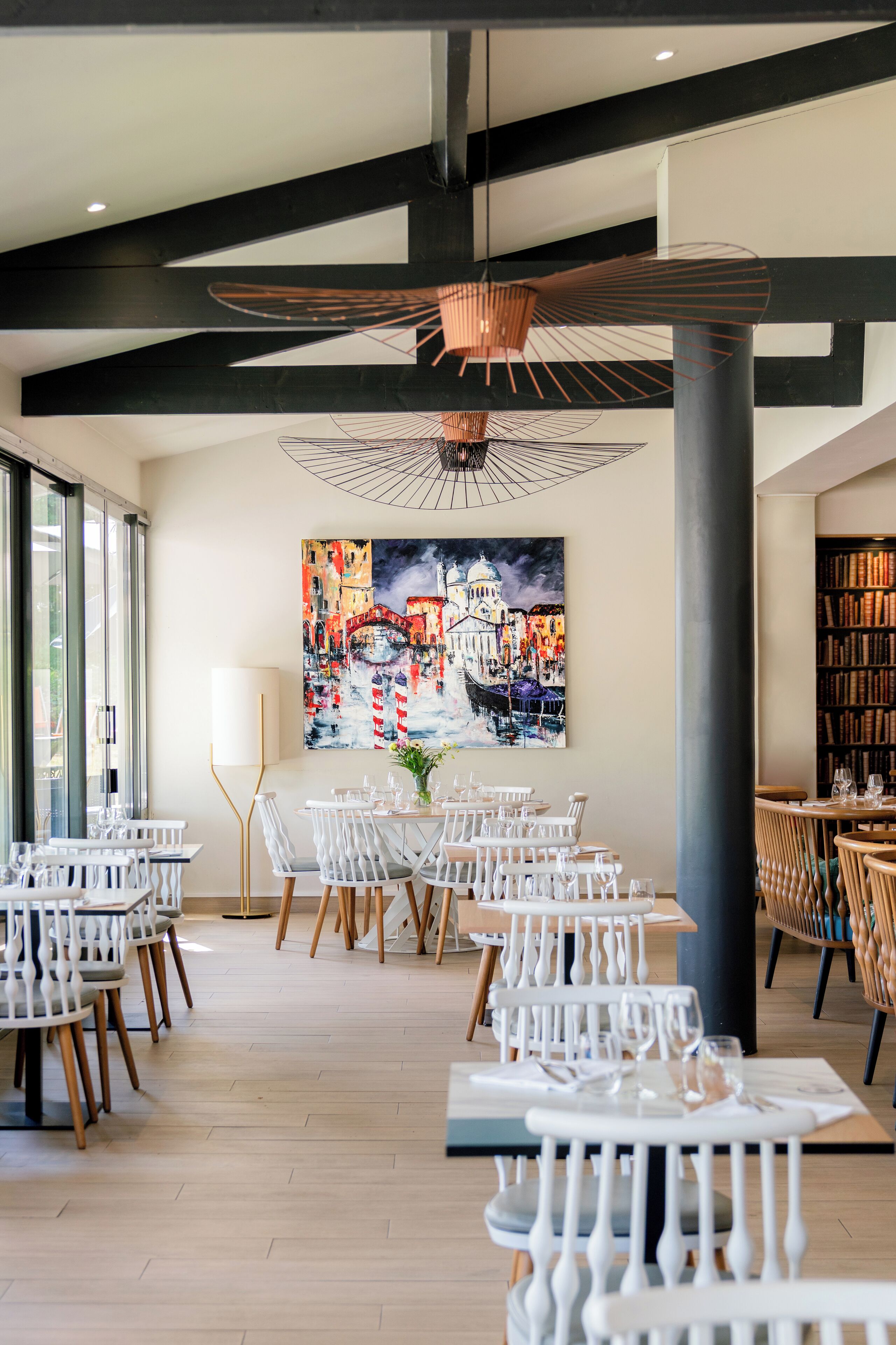 A restaurant with white chairs, tables, and a large painting of a cityscape on the wall.