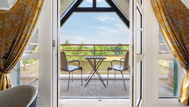 Een kamer met een groot raam, twee stoelen, een tafel en uitzicht op een groen landschap.