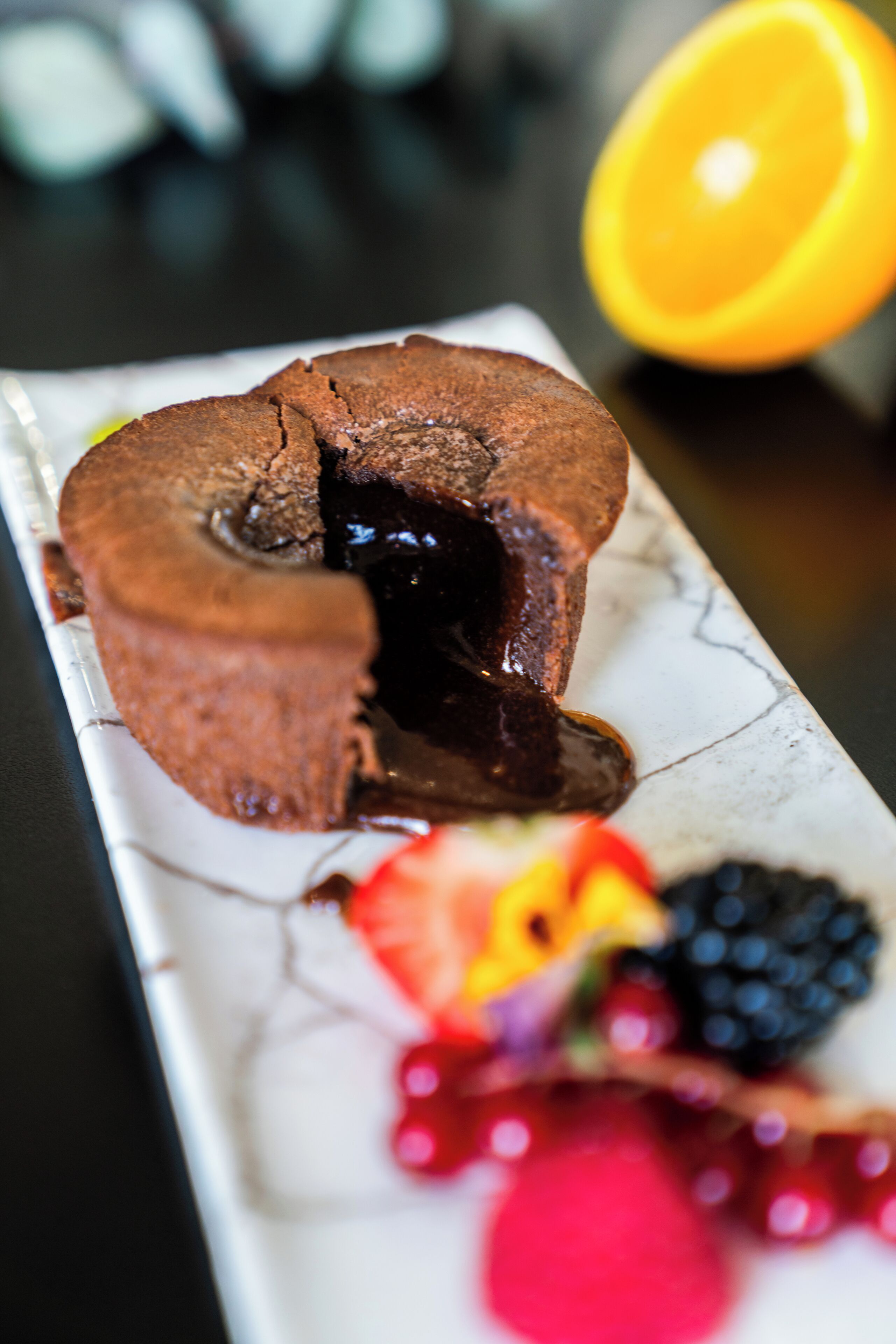Un dessert au chocolat avec une fissure au centre, accompagné de fruits rouges et d’une tranche d’orange, le tout présenté sur une assiette en marbre.