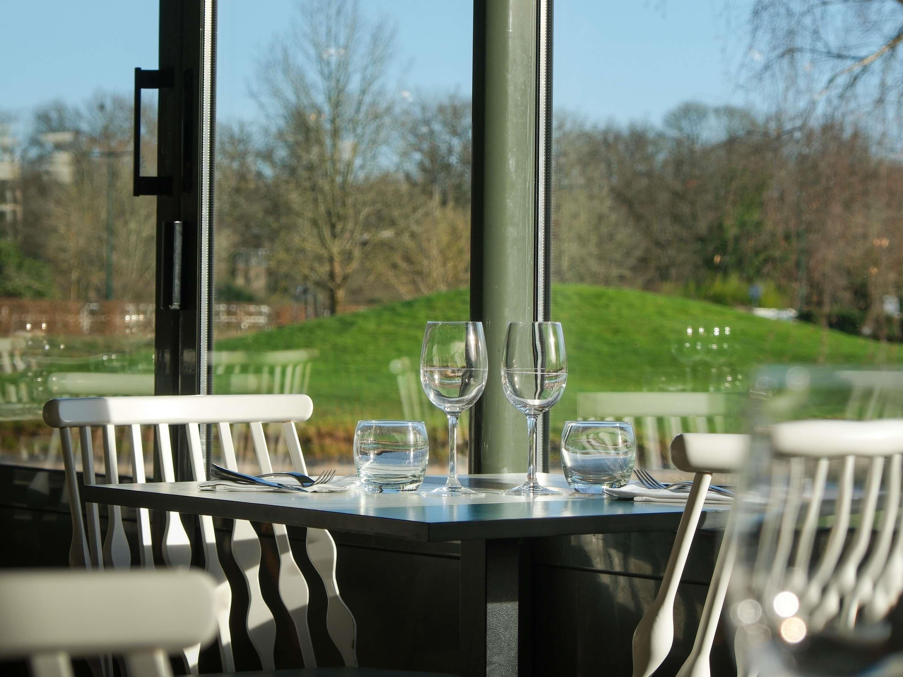A table with four wine glasses and a fork, set against a view of a park with a hill and trees.