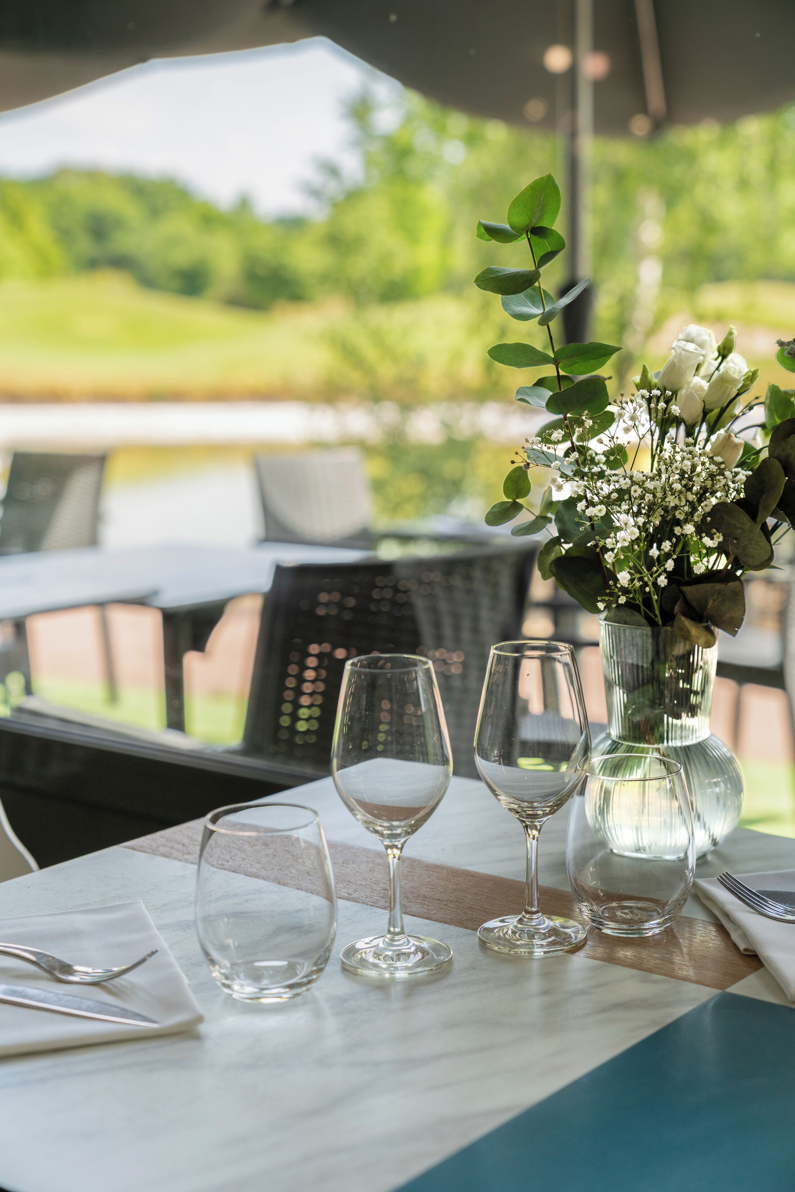 A table set with wine glasses and a vase of flowers, with a view of greenery outside.