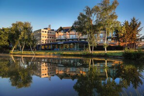 A multi-story hotel with a reflection in the water, surrounded by trees and a clear sky.