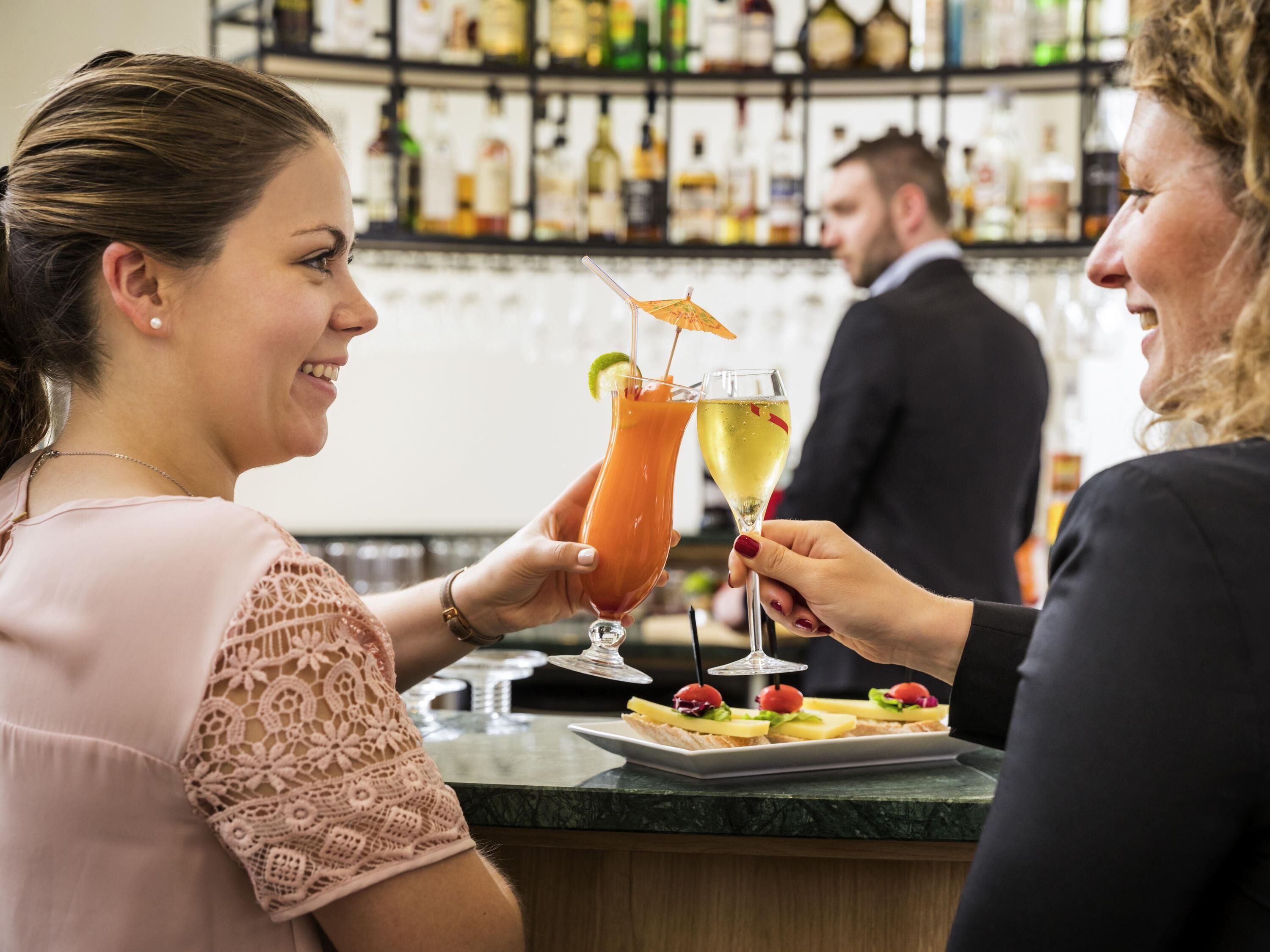 Un bar con mostrador, una persona sosteniendo un cóctel y otra persona con una copa de vino.