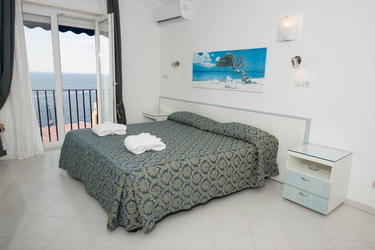 A bedroom with a bed, a nightstand, a glass table, and a painting of a tree by a sea.