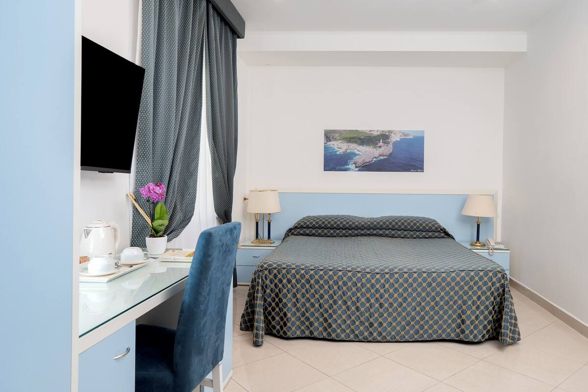A bedroom with a bed, a desk, a chair, a TV, and a painting on the wall.