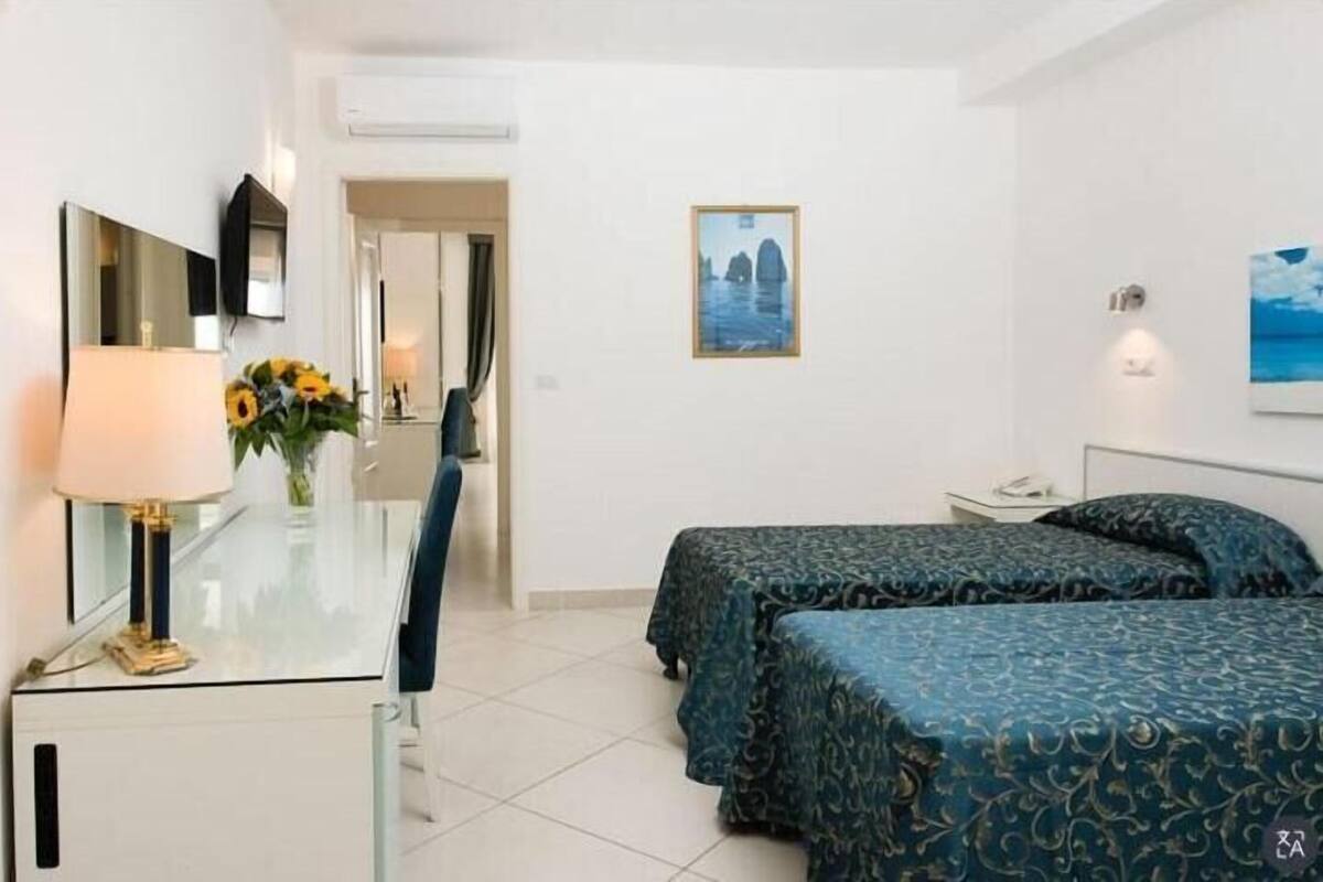 A hotel room with a desk, two beds with patterned bedspreads, a mirror, a vase with sunflowers, and a painting of a coastal scene.
