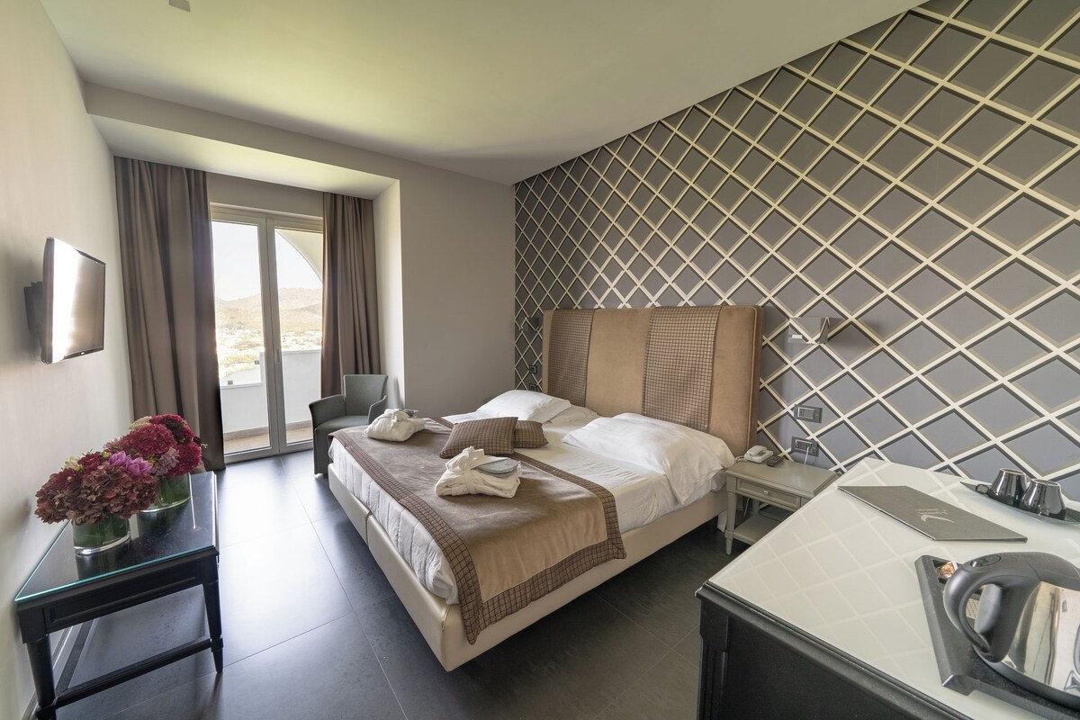 Una habitación de hotel con una cama, un escritorio con teléfono, un espejo y vistas al exterior a través de una puerta corrediza.