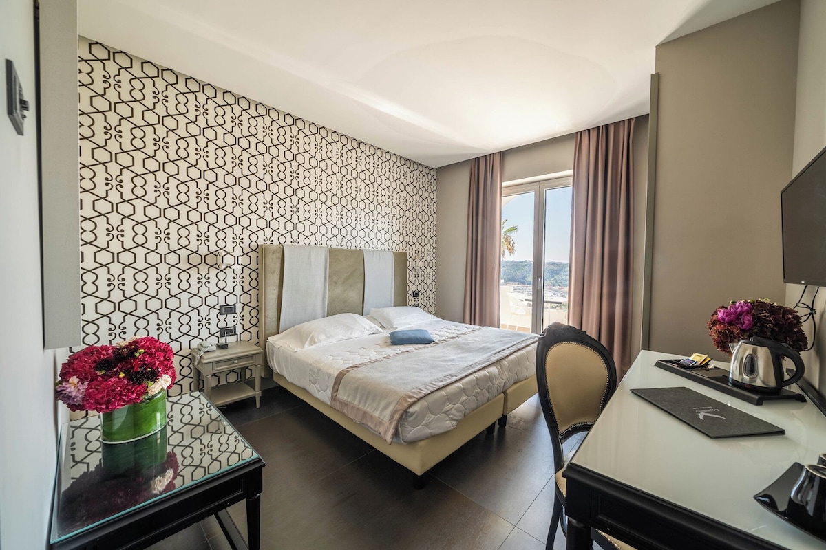 A hotel room with a bed, a desk, a chair, a TV, a window with curtains, and a decorative wall pattern.