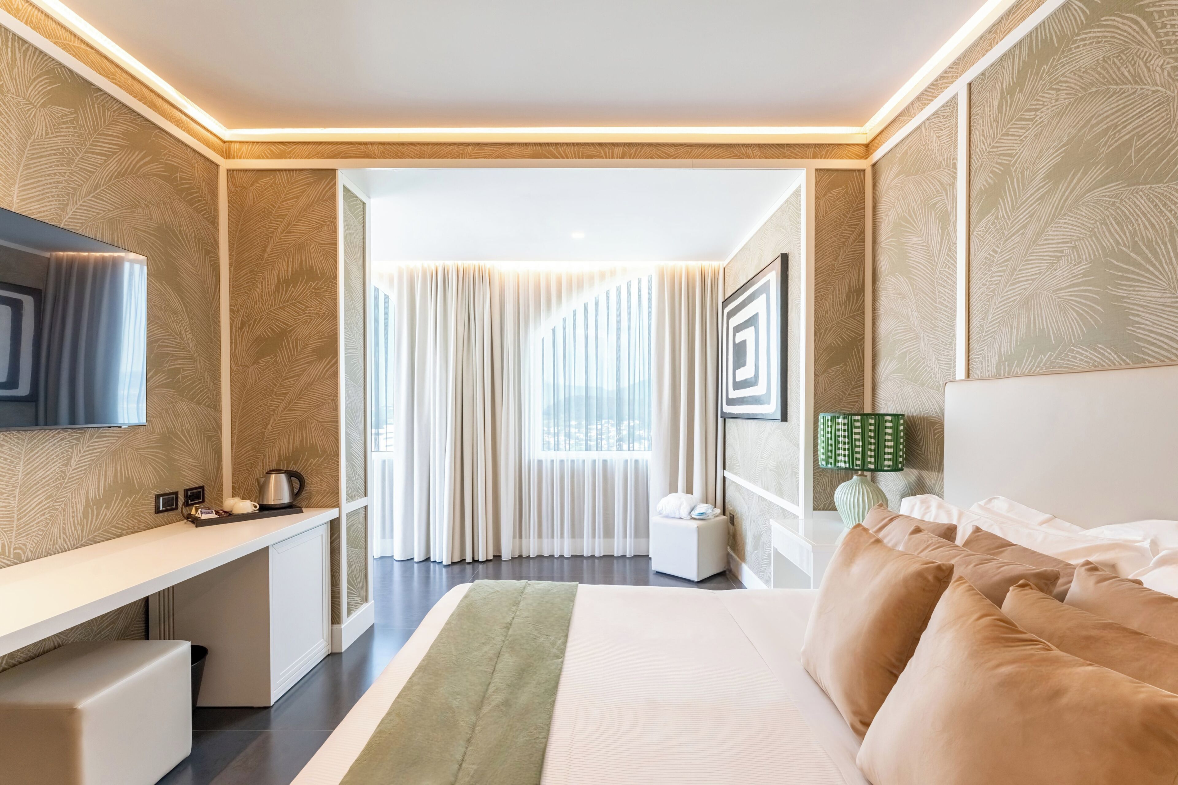 Habitación de hotel con cama, escritorio, televisor, lámpara y vistas a la ciudad a través de amplios ventanales.