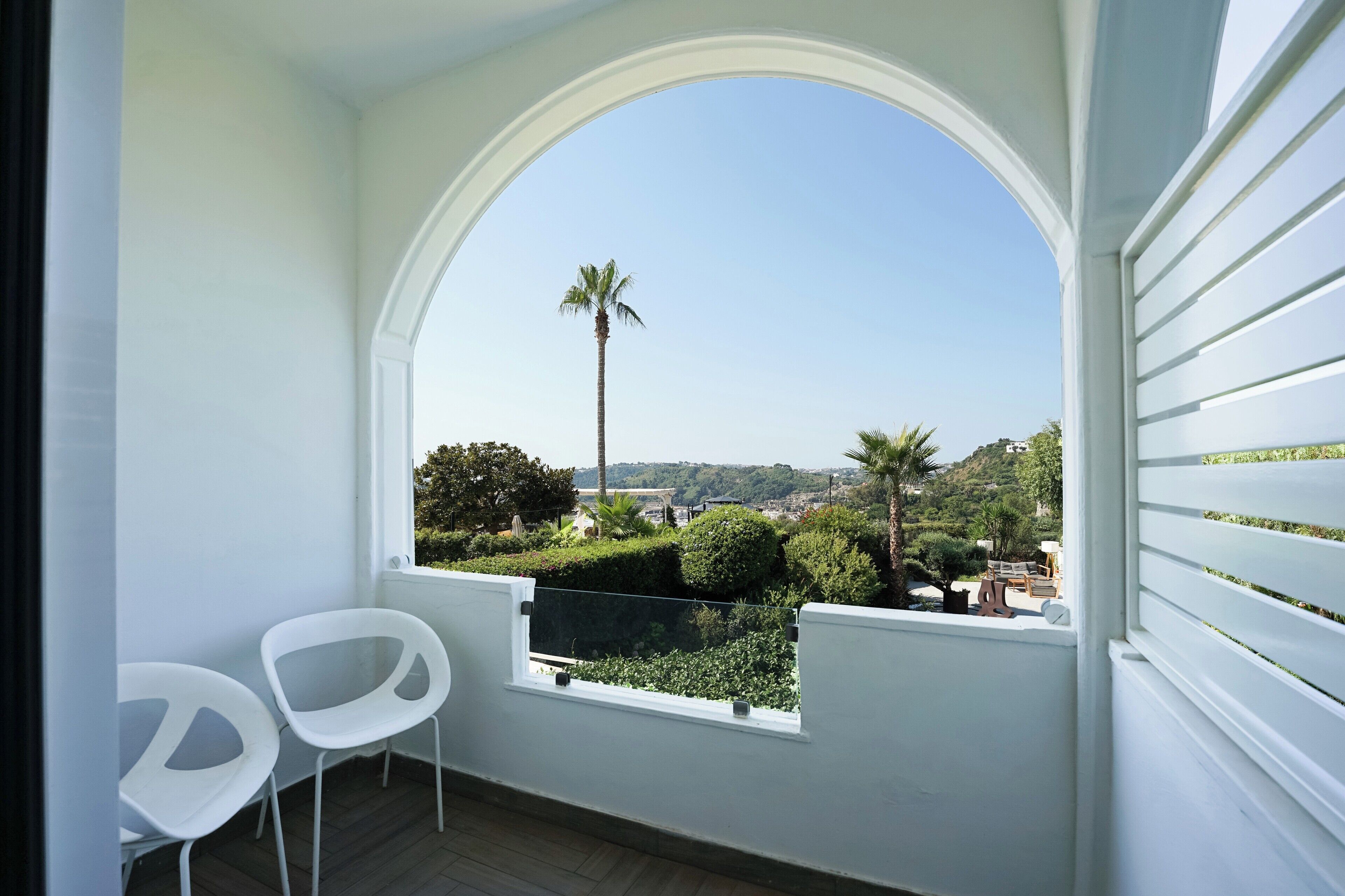 Un balcón con barandilla blanca, dos sillas blancas y una ventana que da a un paisaje verde con árboles y una palmera.