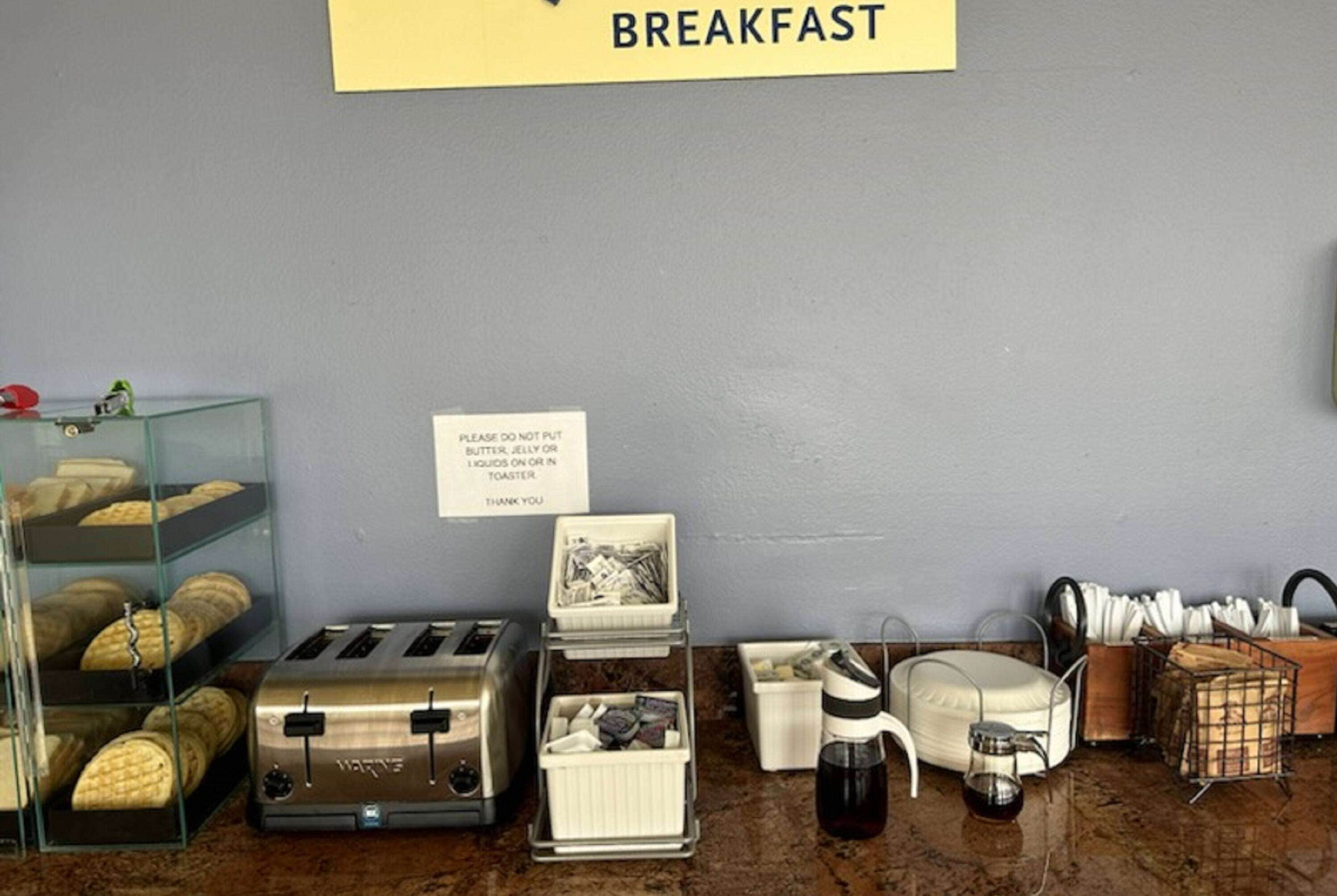 Un área de desayuno con un cartel que indica 'BREAKFAST', un tostador, una cafetera y un letrero que advierte no colocar mantequilla, jalea ni líquidos sobre el tostador.