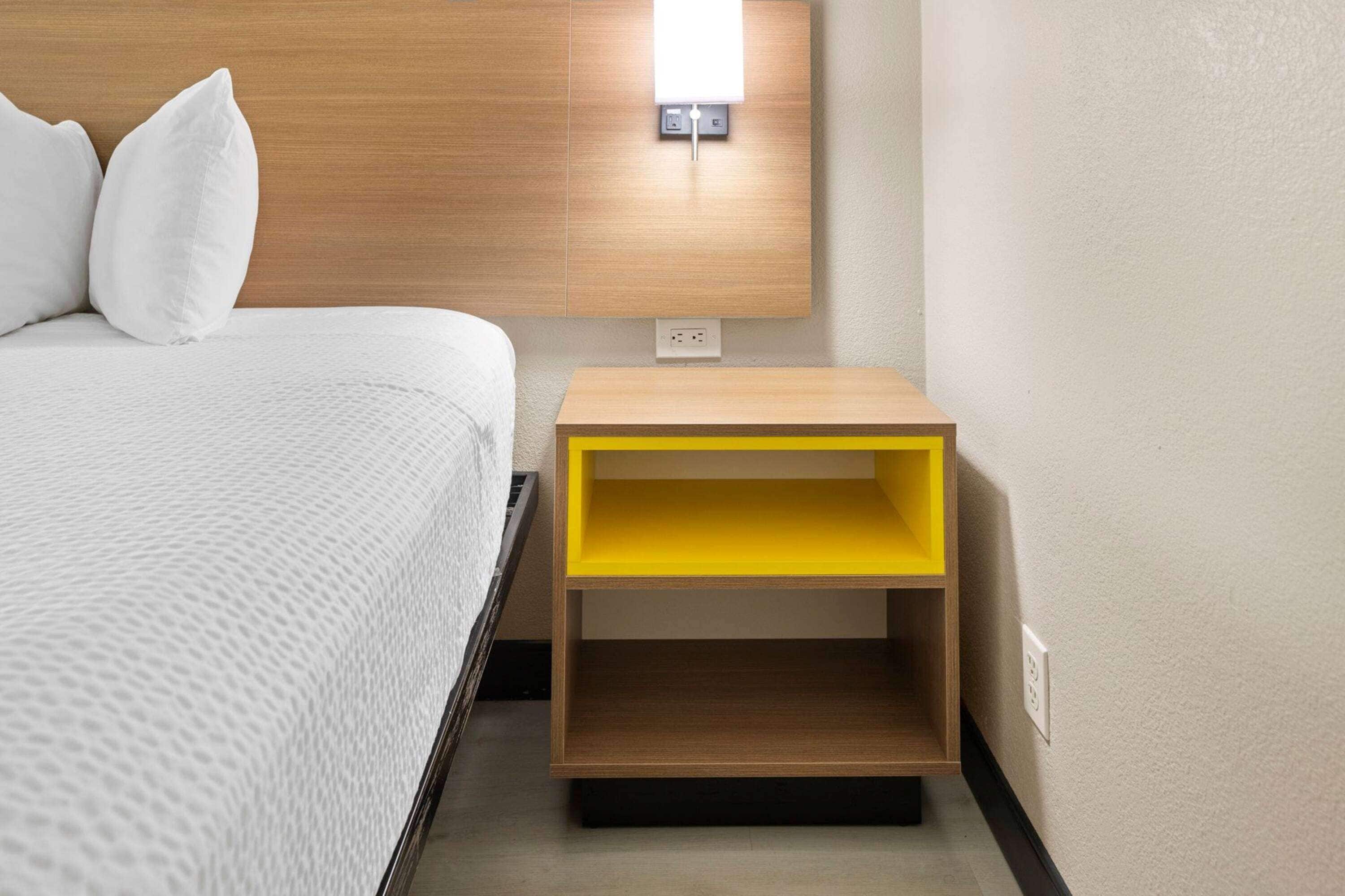 A neatly made bed with white bedding and pillows, a wooden headboard, and a small yellow nightstand with a power outlet.