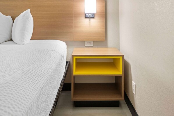 A neatly made bed with white bedding and pillows, a wooden headboard, and a small yellow nightstand with a power outlet.