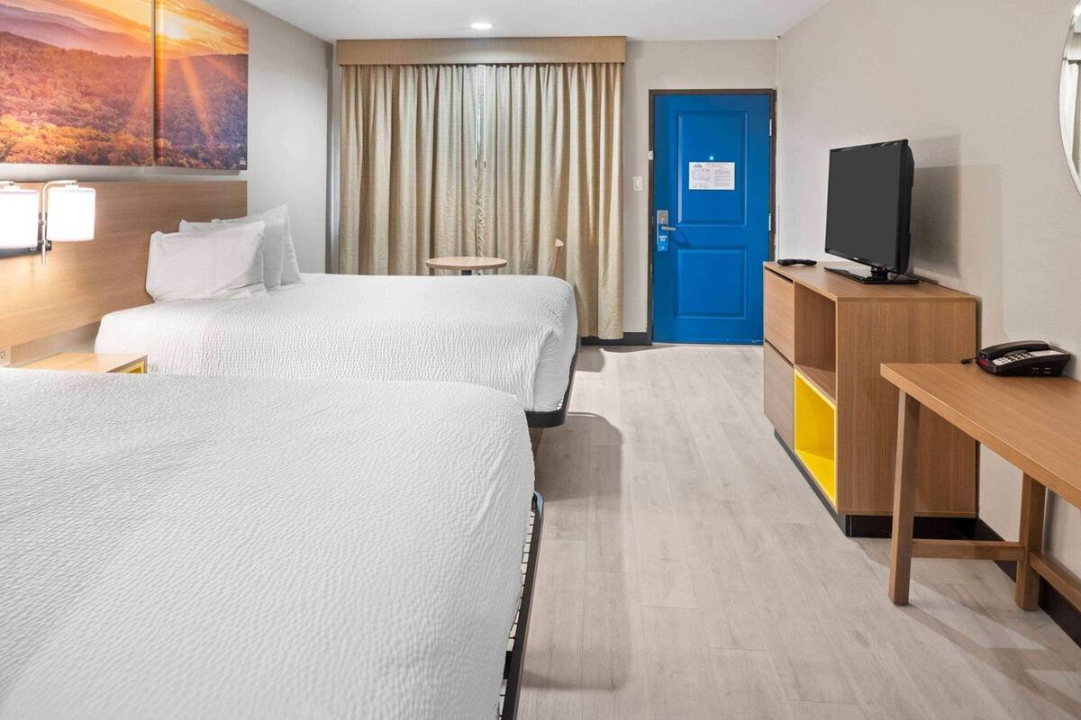 Ein Hotelzimmer mit zwei Betten, einem Fernseher, einem Schreibtisch, einem kleinen Tisch, einer blauen Tür und einem Bild an der Wand.