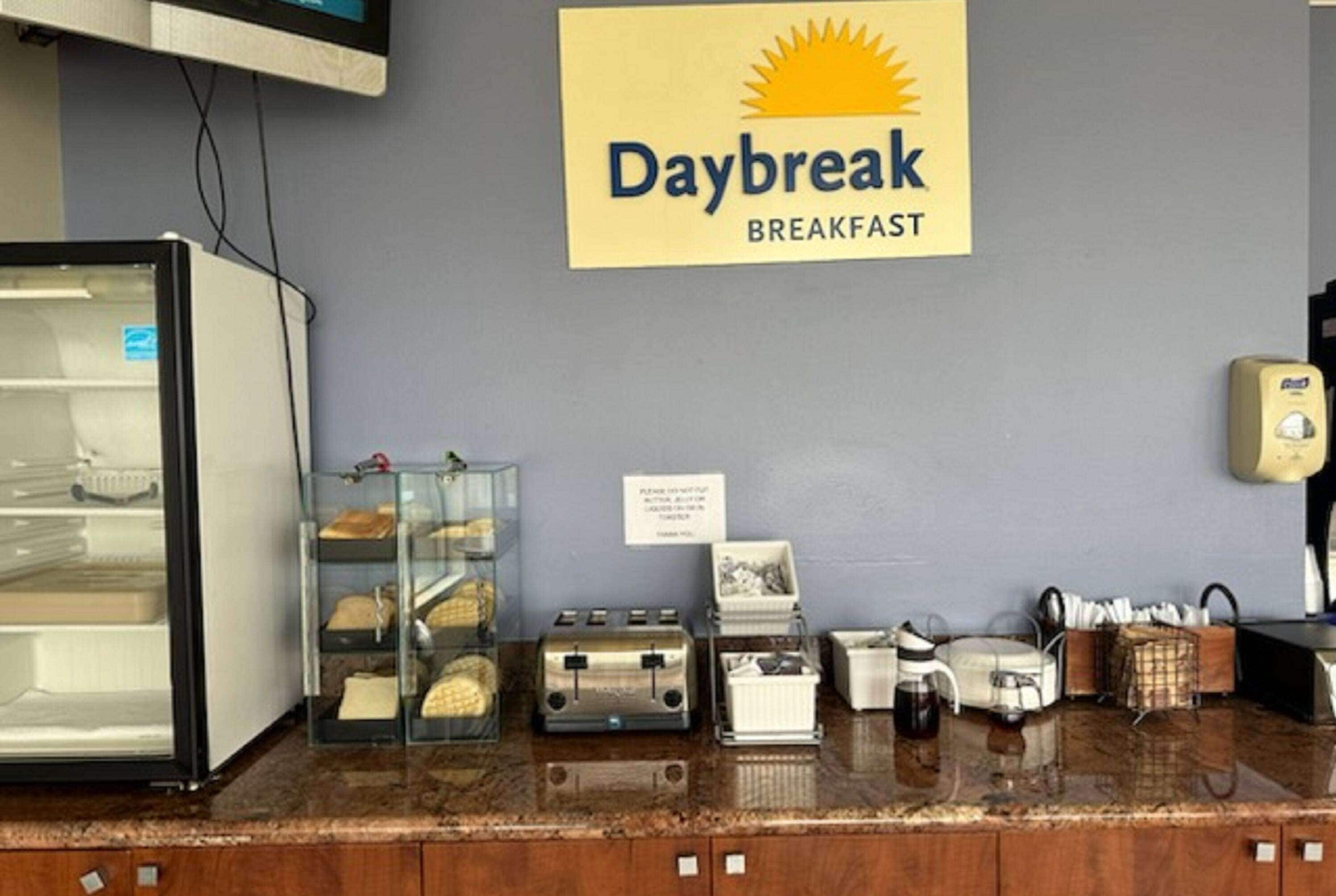 Una barra de desayuno con un letrero que dice "Daybreak Breakfast", un refrigerador, una vitrina de vidrio con productos de repostería, una tostadora y una cafetera.