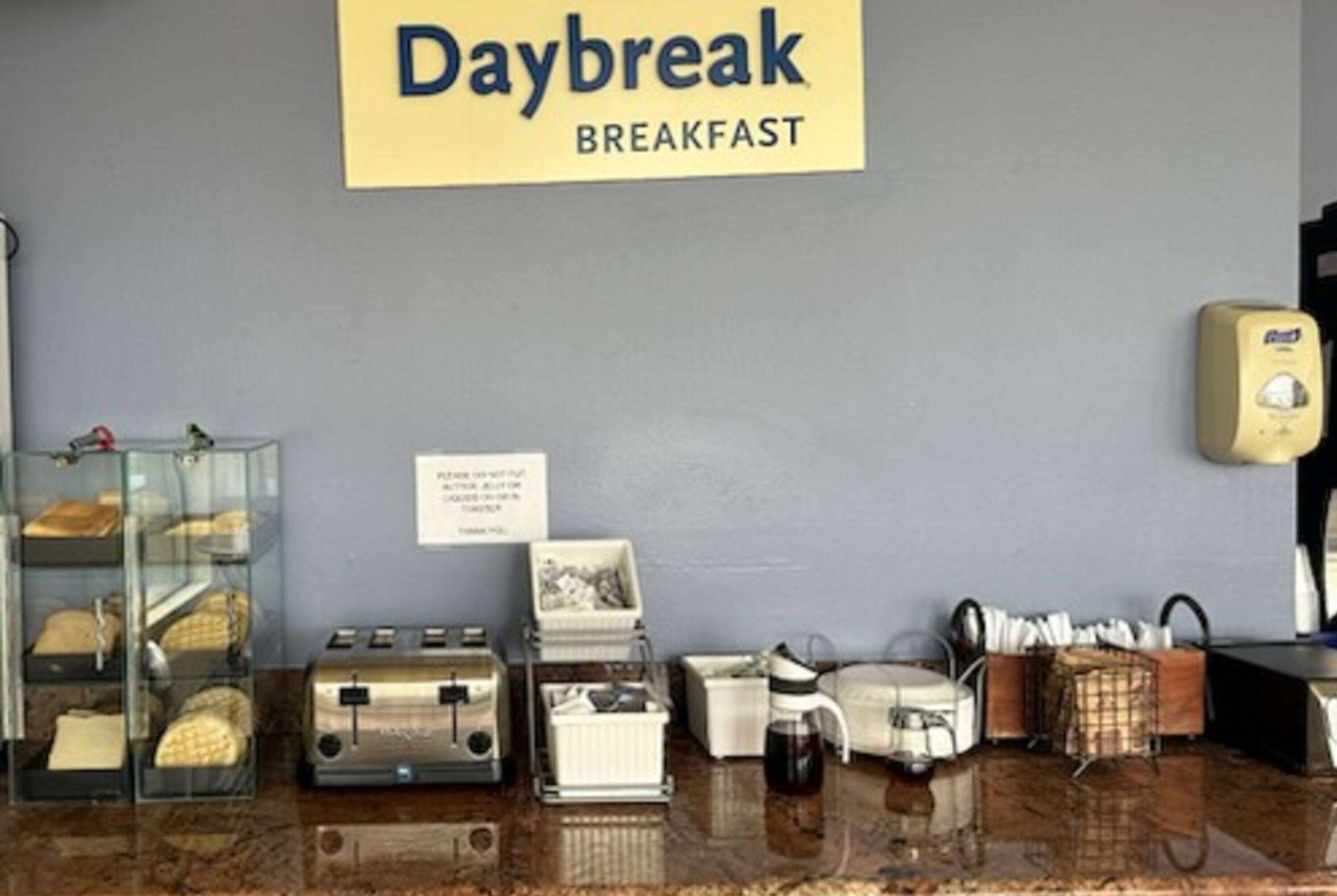 A breakfast counter with a sign reading "Daybreak Breakfast," a coffee machine, a toaster, and a display case with pastries.