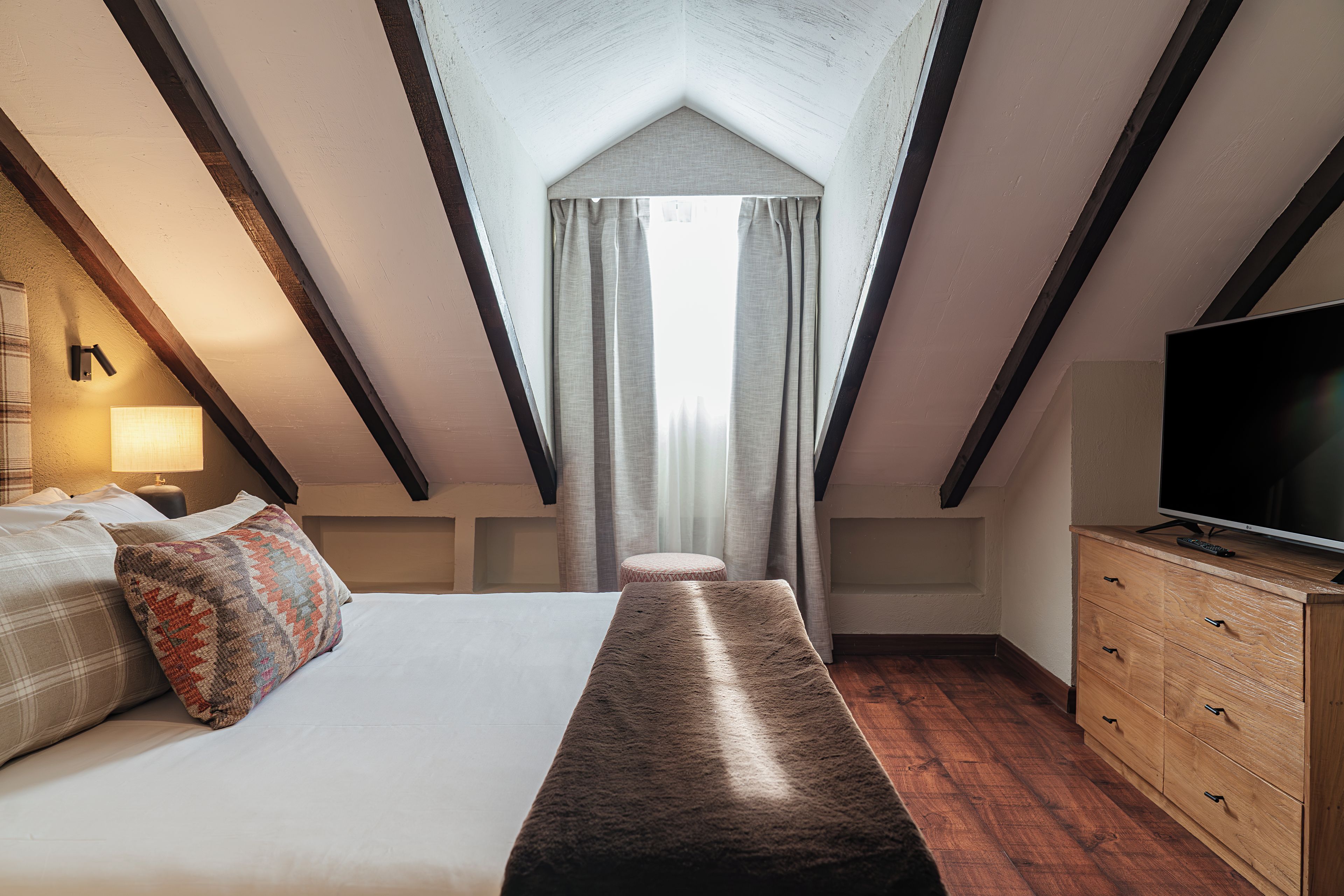 A bedroom with a sloped ceiling, a bed, a bench, a TV, and a dresser.