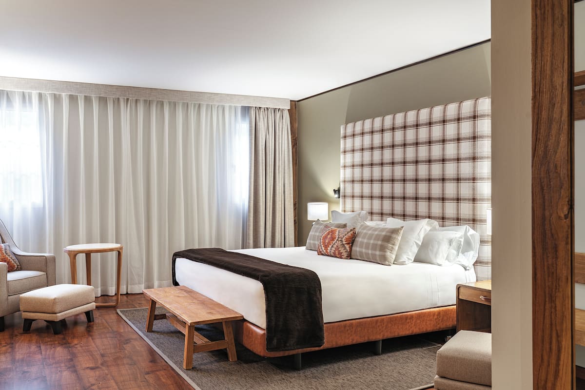 Una habitación de hotel moderna con una cama grande, un banco de madera, una mesita pequeña y un sofá.