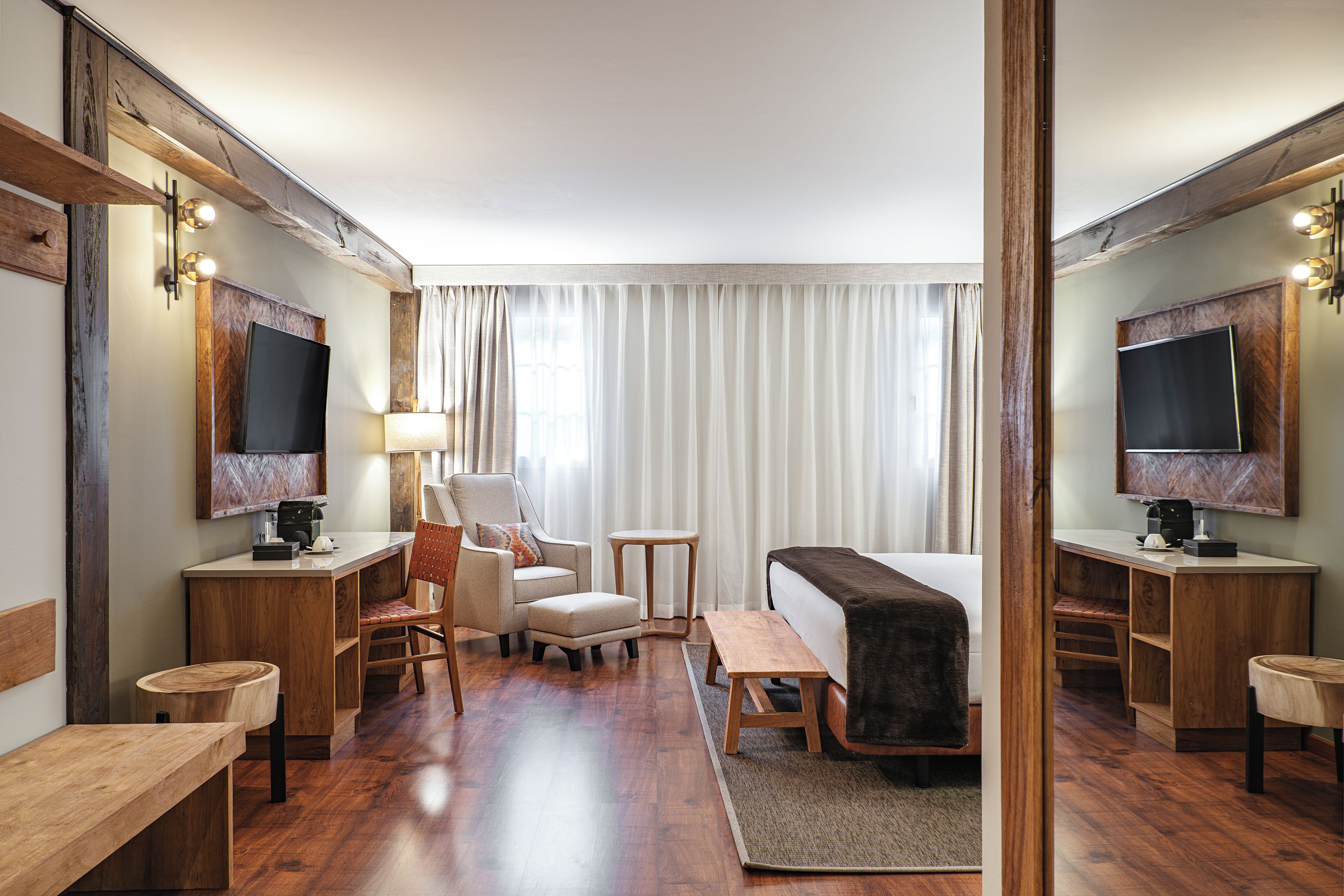Habitación de hotel con piso de madera, una cama con manta oscura, un escritorio con computadora, una silla, una mesita y un televisor sobre un pedestal de madera.