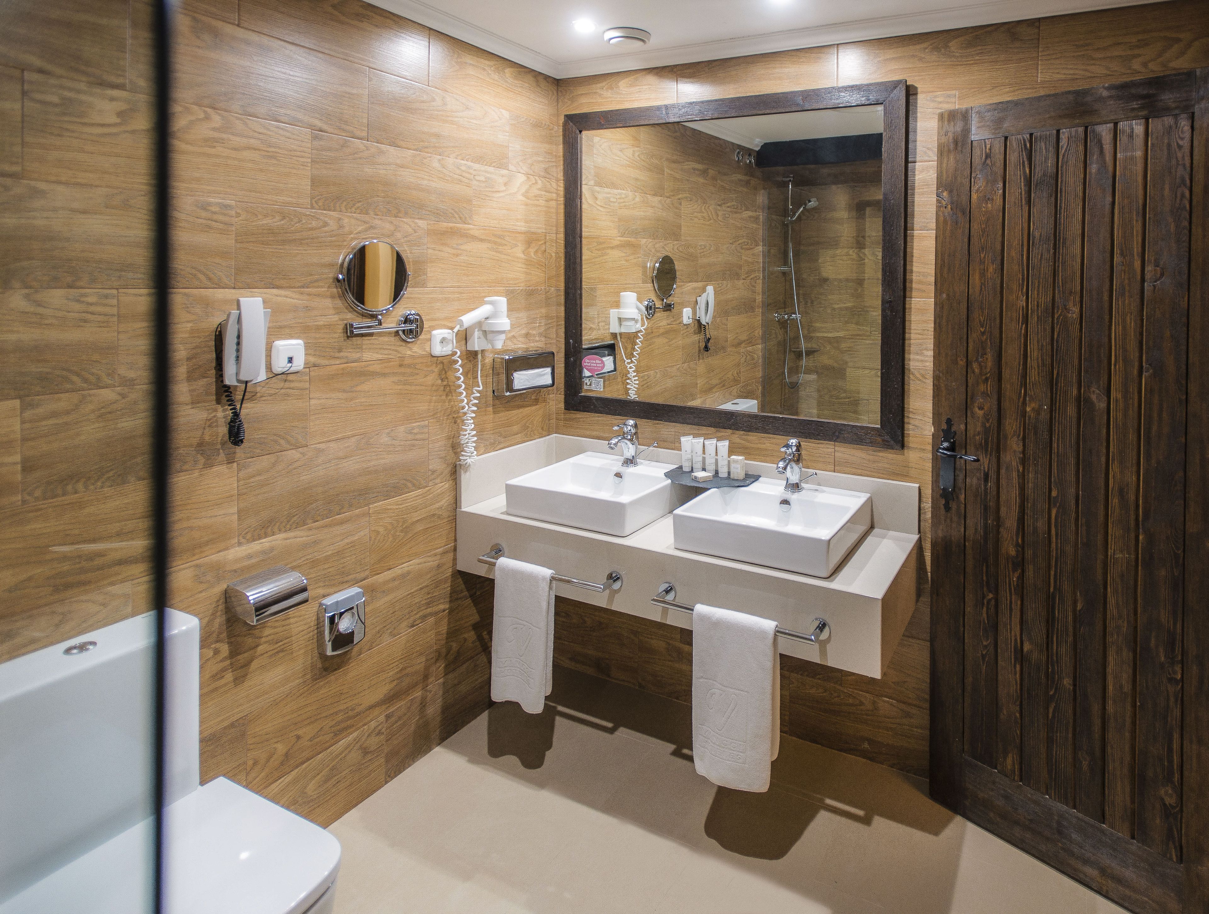 A bathroom with wooden walls, two sinks, a large mirror, a shower, and a toilet.