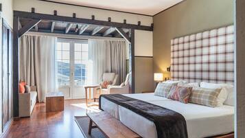 A hotel room with a large bed, a wooden bench, a small table, and a chair. There is a window with curtains and a view of the outdoors.