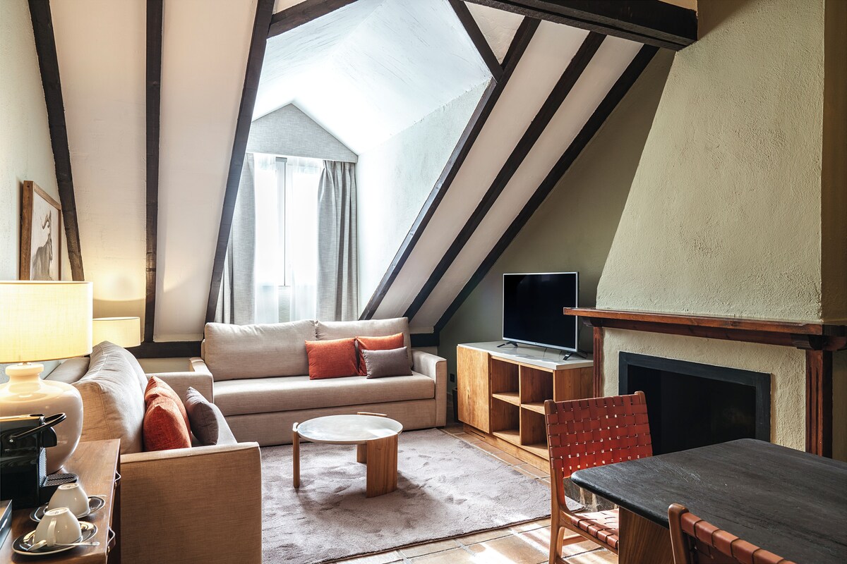 Una sala de estar estilo loft acogedora, con techo alto, vigas a la vista, chimenea, sofá, una mesa de centro y televisor.