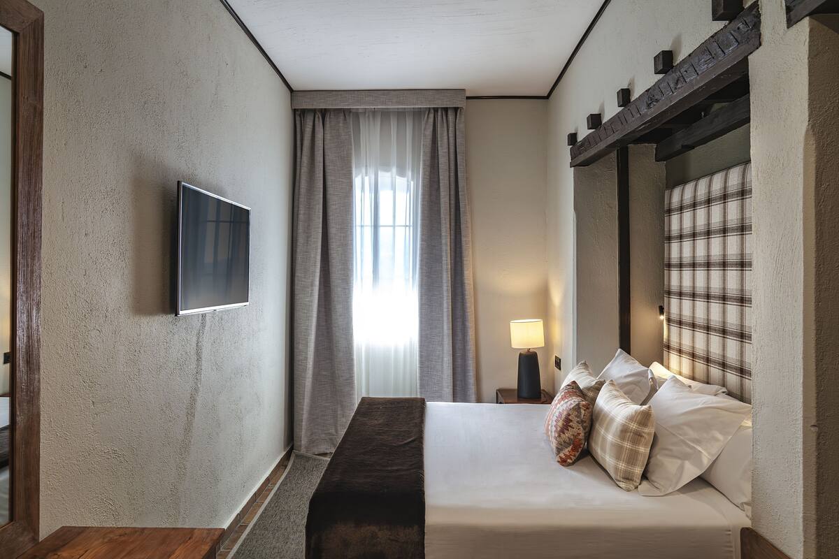 Habitación de hotel con cama, televisor montado en la pared, ventana con cortinas, mesita de noche con lámpara y cabecera con estampado de cuadros.