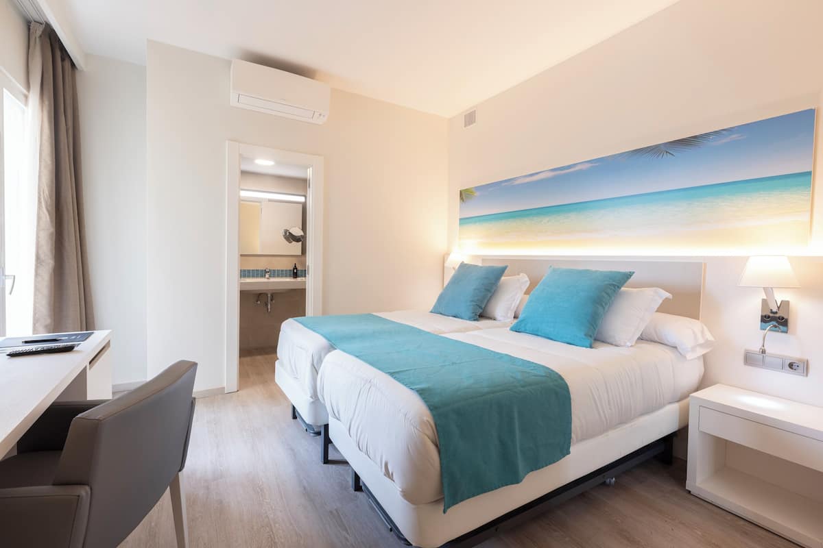 Une chambre d’hôtel comprenant un lit, un bureau, une chaise, une table de chevet et un tableau représentant une scène de plage.