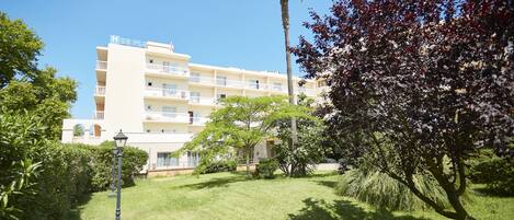 Ein Hotel mit heller Fassade, mehreren Balkonen und einem gepflegten Rasen mit einer Palme und einem blühenden Baum.