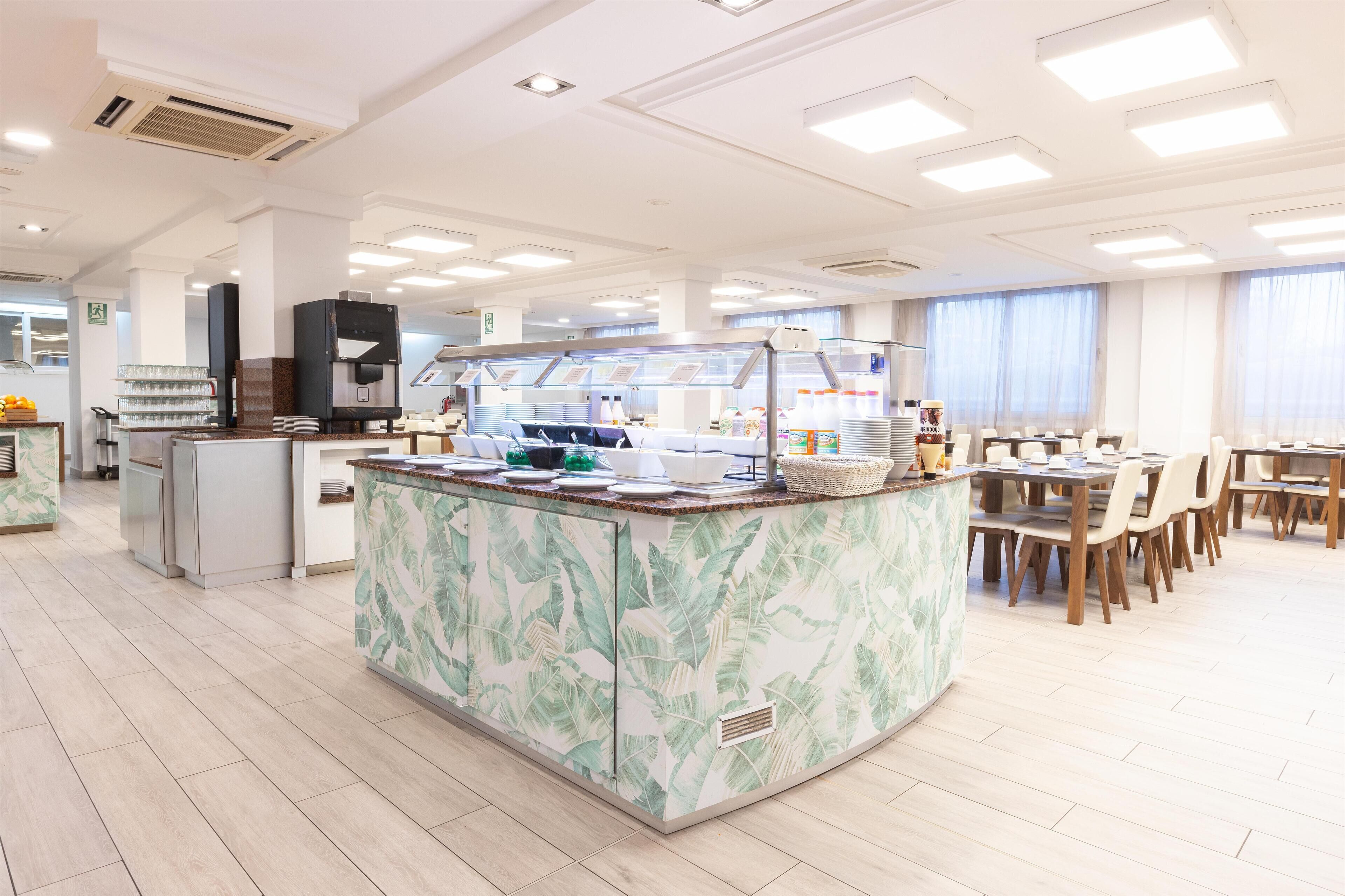 A spacious dining area with multiple tables and chairs, a central buffet station with a coffee machine, and a kitchen area in the background.