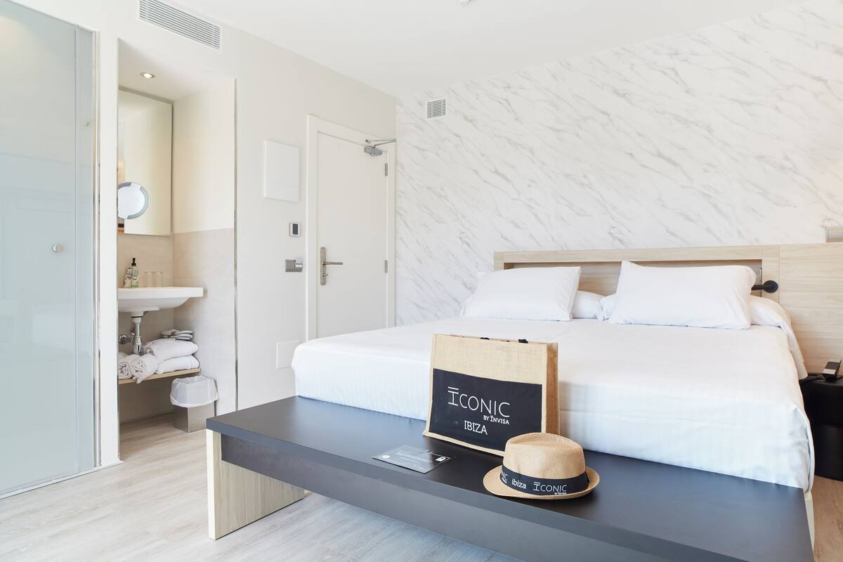 Une chambre d’hôtel moderne avec un lit bien fait, une table de chevet sur laquelle se trouvent un chapeau et un panneau, et une salle de bain équipée d’un lavabo et de serviettes.