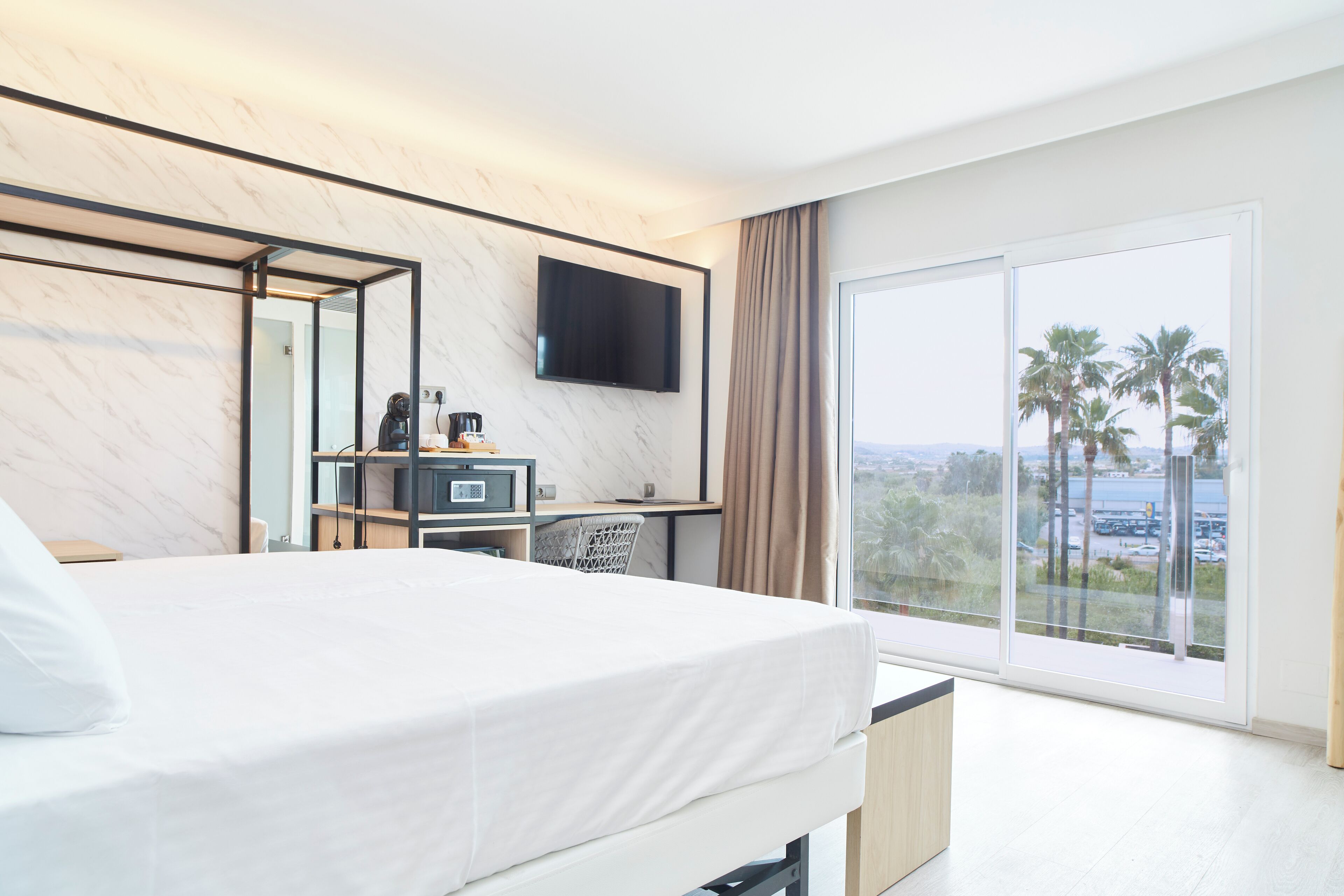 A hotel room with a bed, a desk with a flat-screen TV, a coffee machine, and a large window with a view of palm trees and a body of water.