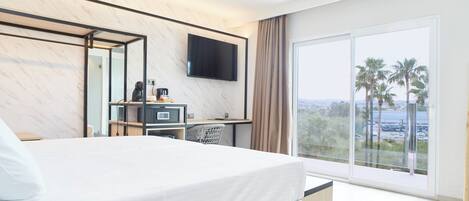 Habitación de hotel con cama, escritorio con televisor de pantalla plana, cafetera y ventana grande con vista a palmeras y una masa de agua.