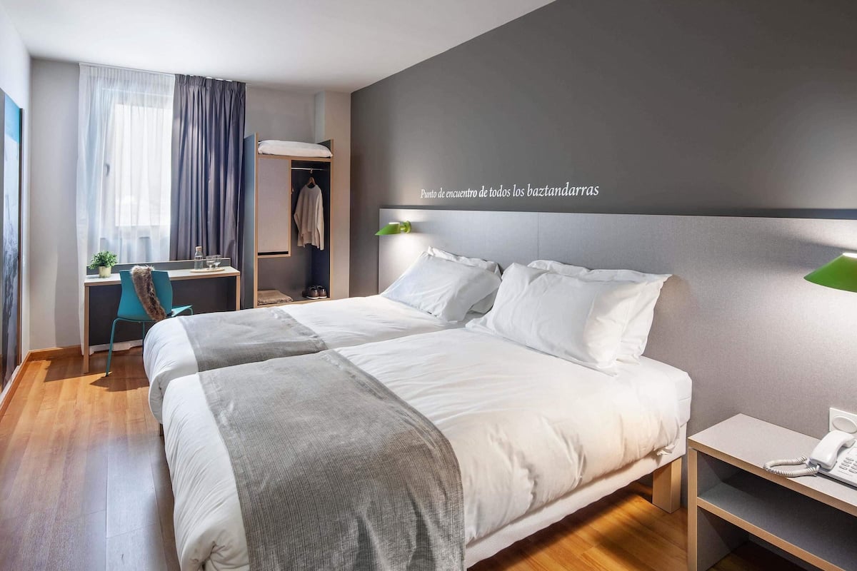 A modern hotel room with a large bed, a desk, a chair, and a wardrobe.